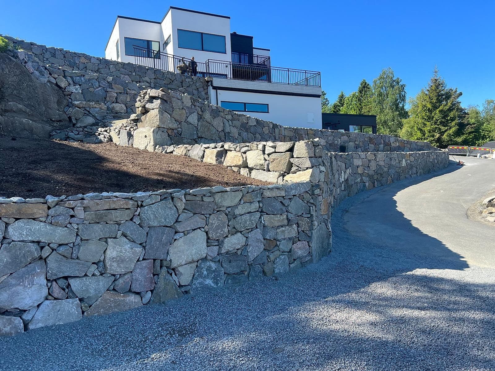 Nybyggda murar av natursten i flera nivåer längs en infart, framför ett modernt hus.