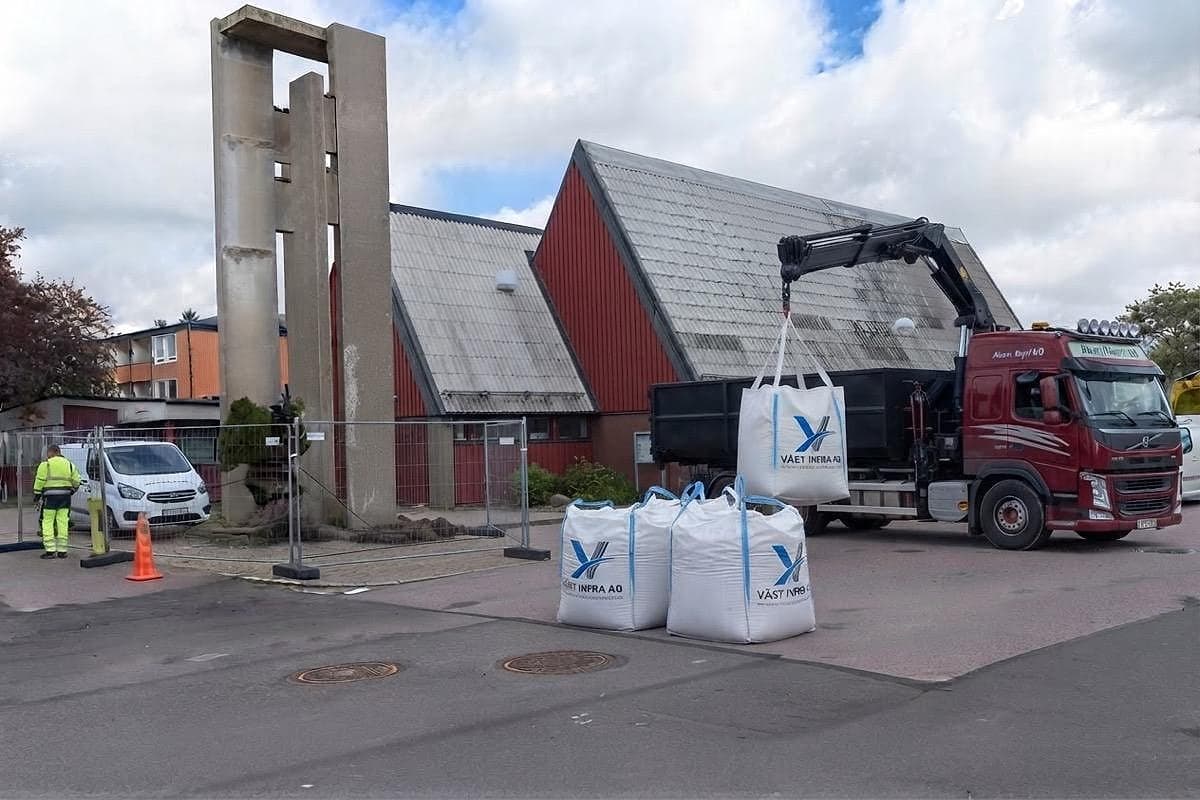 Kranbil lyfter storsäckar märkta Väst Infra AB utanför en kyrka.